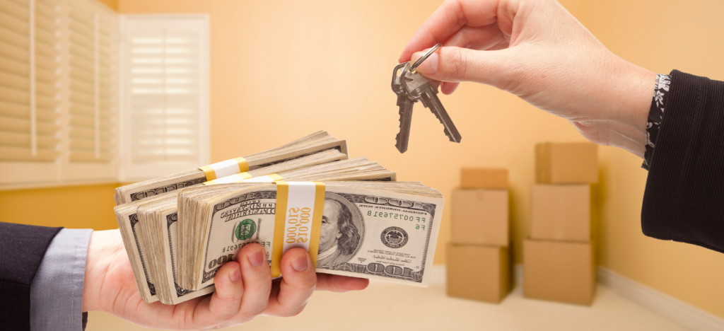 A close-up of a hand exchanging a set of house keys for a large stack of hundred-dollar bills, with moving boxes in the background, symbolizing a fast cash home sale.