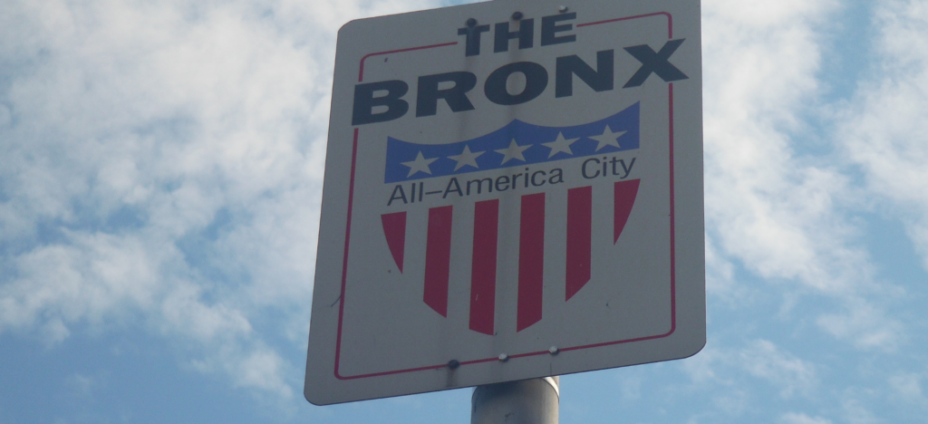 A street sign reading “The Bronx All-America City” with a patriotic red, white, and blue shield, set against a partly cloudy sky, representing the Bronx community.
