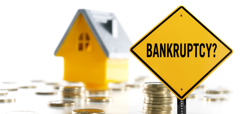 A yellow traffic sign with the word "Bankruptcy" stands next to a model house and stacks of coins, symbolizing financial challenges and the need for solutions during bankruptcy.