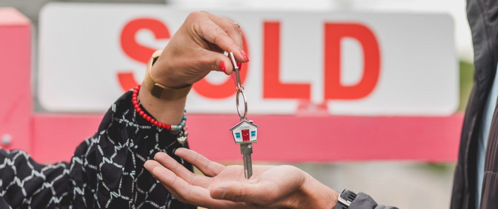 Close-up of one person handing over a house-shaped keychain with a key to another person's open palm, with a red 'SOLD' sign in the background, symbolizing a successful home sale in New Jersey or New York