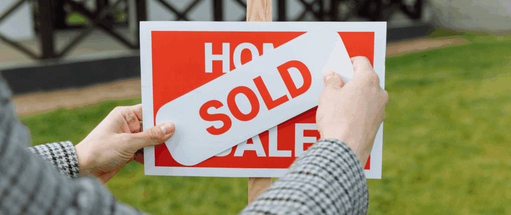Close-up of a person placing a “SOLD” sticker over a red “Home For Sale” sign in front of a house, symbolizing a successful and stress-free home sale.