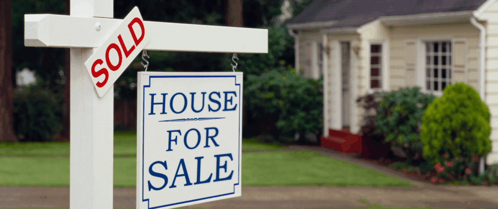 A white “House For Sale” sign with a red “SOLD” label in front of a suburban home, representing a quick and successful cash home sale.