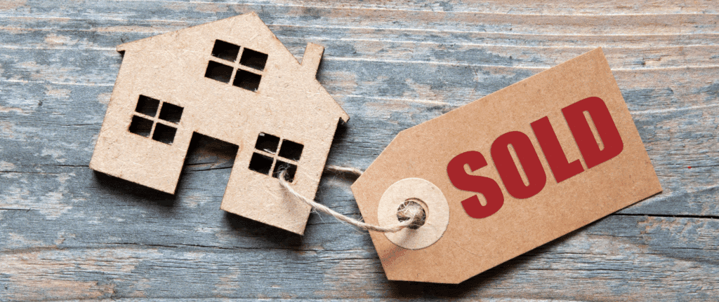 A cardboard cutout of a house attached to a brown tag labeled "SOLD," resting on a rustic wooden surface, representing a fast and easy home sale in Dutchess County.