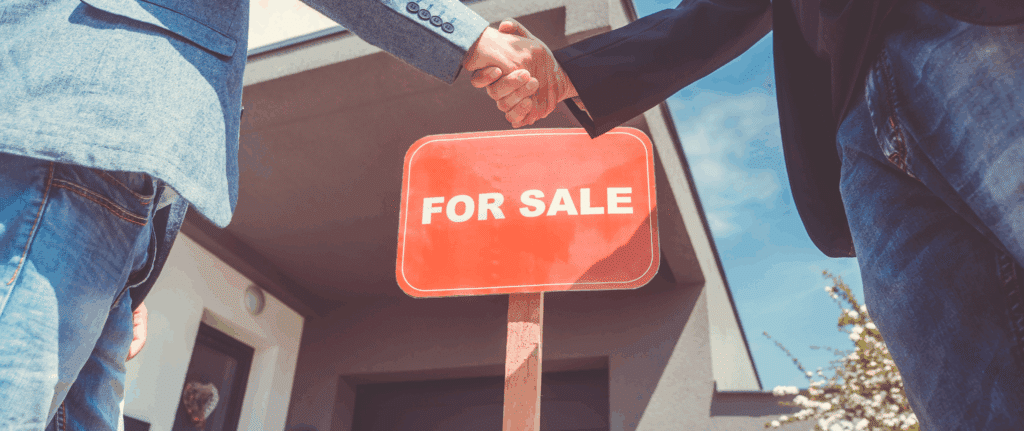 Two people in business attire shaking hands in front of a red "For Sale" sign outside a modern home, symbolizing a successful cash home sale in New York.